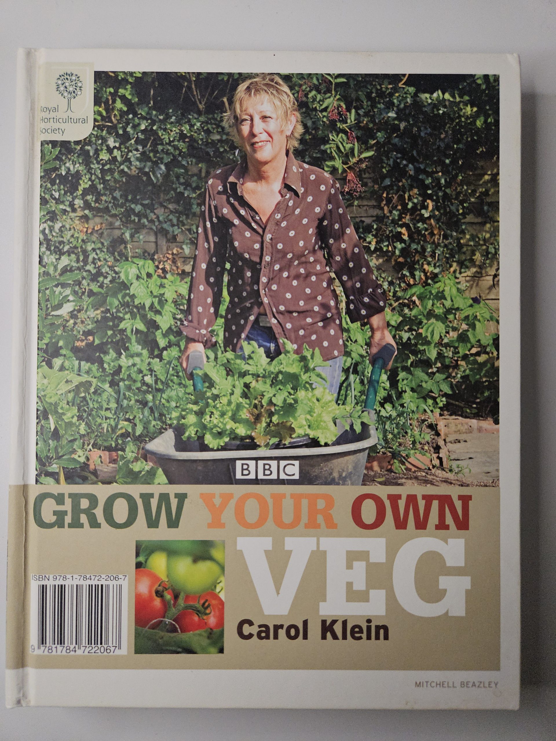 A woman standing in a garden with plants in a wheelbarrow, wearing a brown polka-dot shirt; BBC logo visible and book title Grow Your Own Veg.